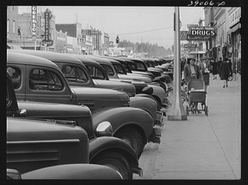 A historic street scene with rows of vintage cars and a woman pushing a stroller along the sidewalk.