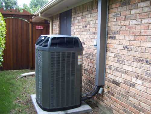 An outdoor air conditioning unit installed next to a brick wall, with greenery in the background.