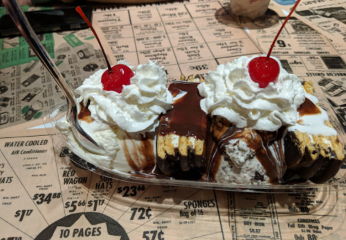 A large ice cream sundae with whipped cream and cherries on top, served in a glass dish on a vintage menu backdrop.