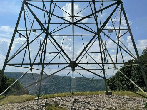A large power line tower stands against a backdrop of rolling hills and a cloudy sky.