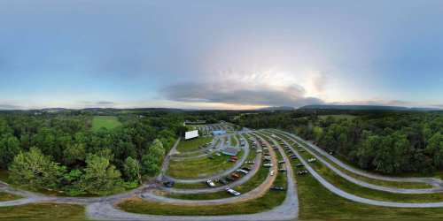 Aerial view of a drive-in movie theater surrounded by trees and fields, with parked cars and a cloudy sky.
