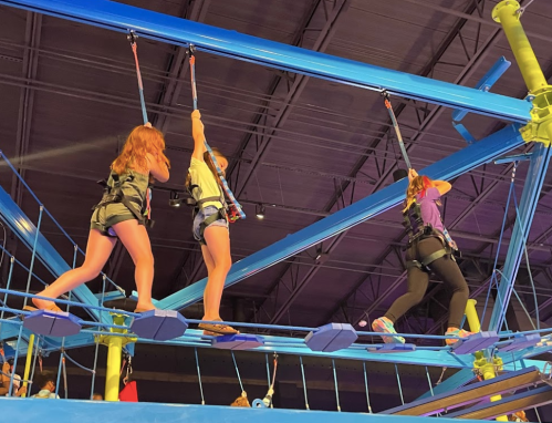 Three children navigate a colorful indoor ropes course, secured with harnesses, high above the ground.