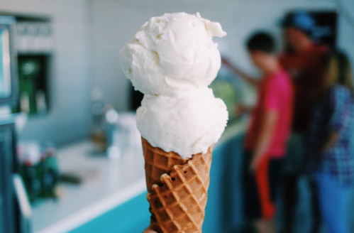 A close-up of a double scoop of vanilla ice cream on a waffle cone, with people in the background.