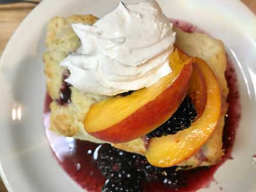 A biscuit topped with whipped cream, sliced peaches, and blackberry sauce on a white plate.