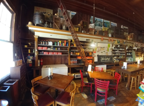 Interior of a rustic restaurant with wooden tables, colorful chairs, and shelves filled with various food items and decor.