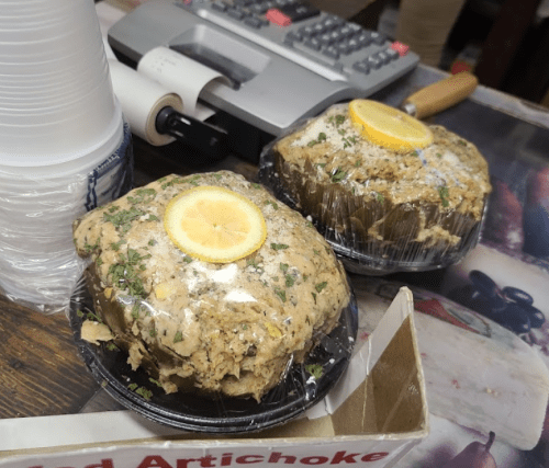 Two wrapped artichoke dishes topped with lemon slices, placed on a counter with a cash register in the background.