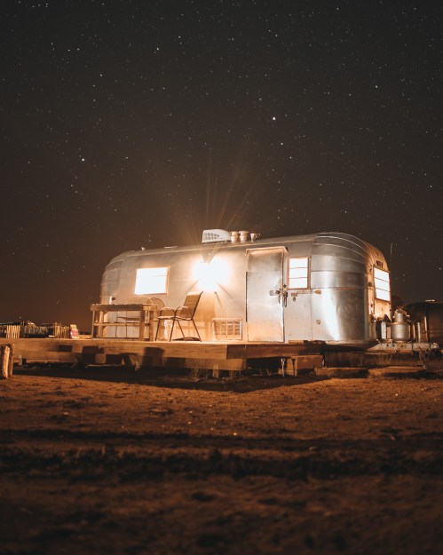 A vintage silver trailer illuminated at night, surrounded by a starry sky and a sandy landscape.