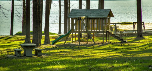 A playground with a slide and wooden structure, surrounded by trees and a lake in the background on a sunny day.