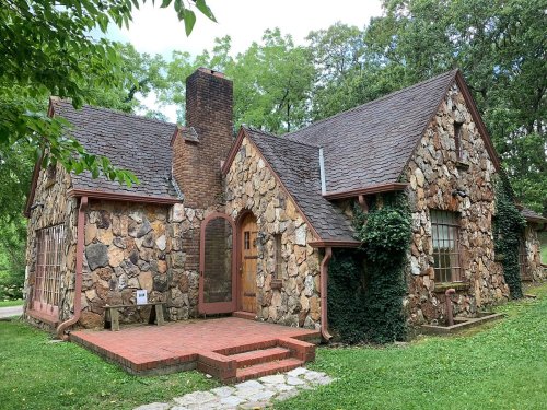 A charming stone cottage with a sloped roof, surrounded by greenery and featuring a brick pathway and wooden door.