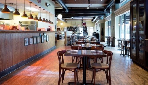 A cozy restaurant interior with wooden tables, chairs, and a warm ambiance, featuring a bar and open kitchen area.