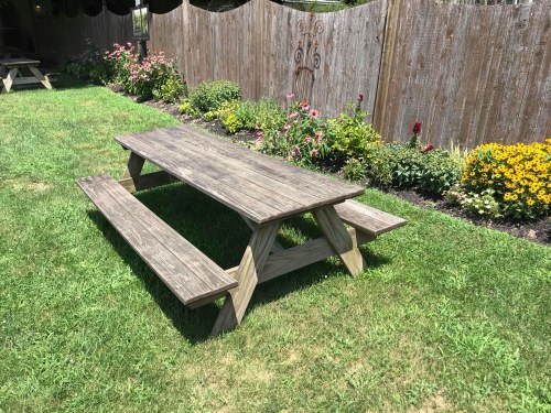 A wooden picnic table on a grassy area surrounded by colorful flowers and a wooden fence.