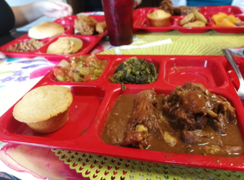A red cafeteria tray with cornbread, meat in gravy, greens, and a salad, set on a colorful tablecloth.