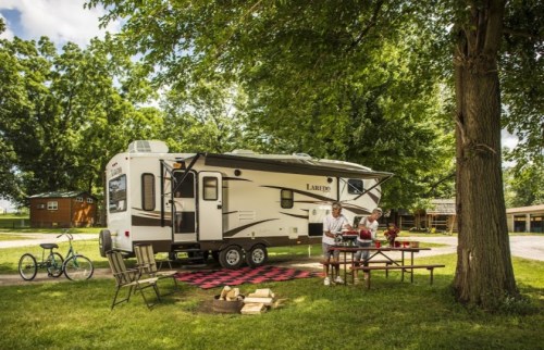 A couple enjoys a picnic outside their RV, surrounded by trees and a grassy area, with a bike nearby.