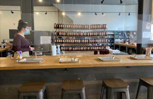 A woman in a mask stands at a wooden bar in a shop with shelves of bottles in the background.