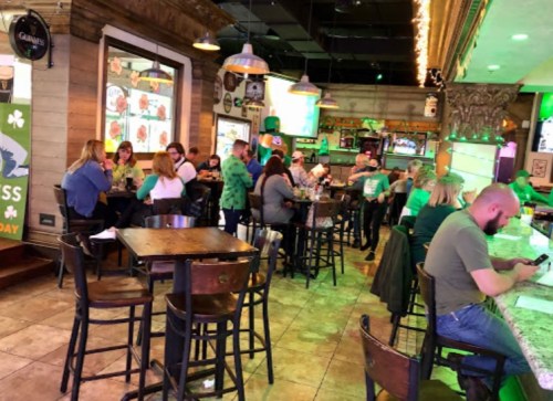 A lively pub scene with patrons in green attire, celebrating St. Patrick's Day, seated at tables and bar stools.