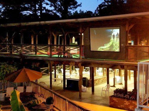 Outdoor area with a large screen showing a movie, surrounded by seating and trees at dusk.