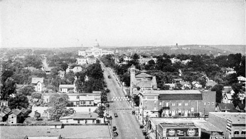 A historic view of a town with a main street, buildings, trees, and a distant government building on a hill.