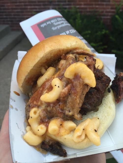 A close-up of a sandwich with pulled meat and macaroni and cheese on a soft bun, held in a hand.