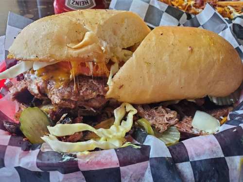 A hearty sandwich filled with meat, cheese, pickles, and onions, served with coleslaw on a checkered paper plate.