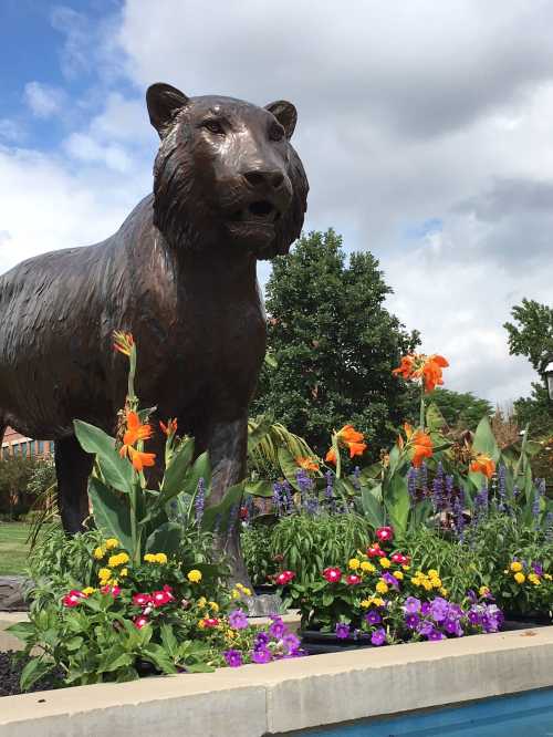 A large bronze bear statue surrounded by colorful flowers and greenery under a partly cloudy sky.