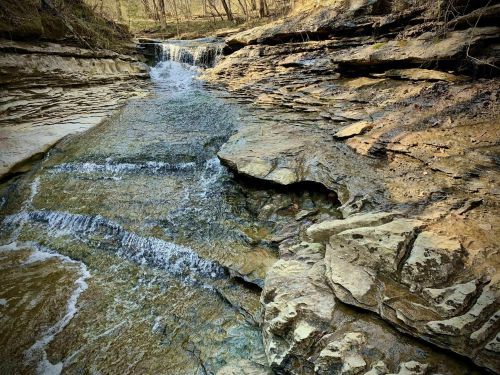 A serene stream flows over rocky terrain, surrounded by trees and natural landscape.