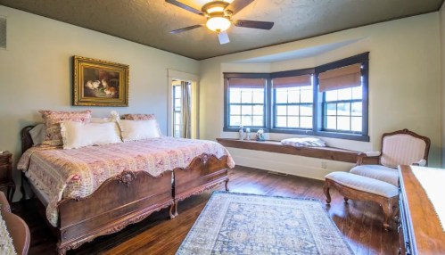 Cozy bedroom with a wooden bed, decorative pillows, bay windows, and a vintage armchair. Warm, inviting atmosphere.
