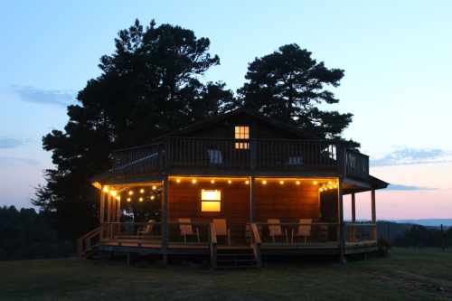 A cozy wooden cabin with string lights, surrounded by trees at dusk, overlooking a scenic landscape.