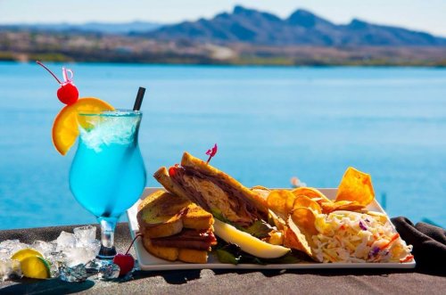 A vibrant blue cocktail beside a plate of pulled pork sandwich, chips, and coleslaw, with a scenic lake view in the background.