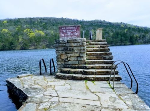 Stone dock with stairs leading into a lake, surrounded by greenery and a warning sign about swimming safety.