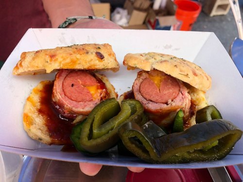 A hand holds a tray with two biscuit sandwiches filled with sausage and cheese, accompanied by sliced green peppers.