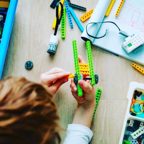 A child builds with colorful LEGO pieces, assembling a vehicle on a wooden floor, surrounded by various LEGO elements.