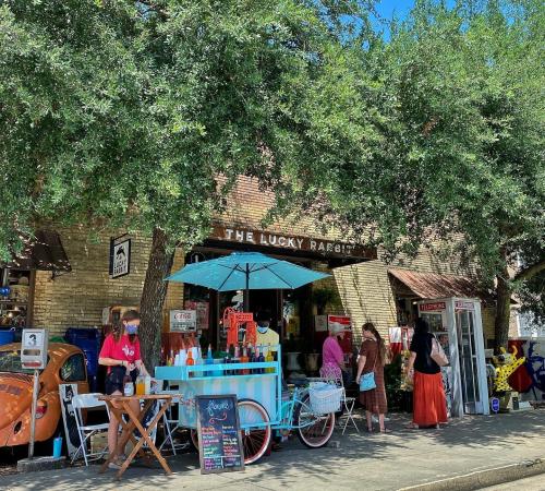 A charming storefront with a blue awning, people browsing outside under leafy trees on a sunny day.