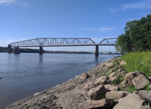 A steel bridge spans a river, with rocky banks and greenery on the shore under a clear blue sky.