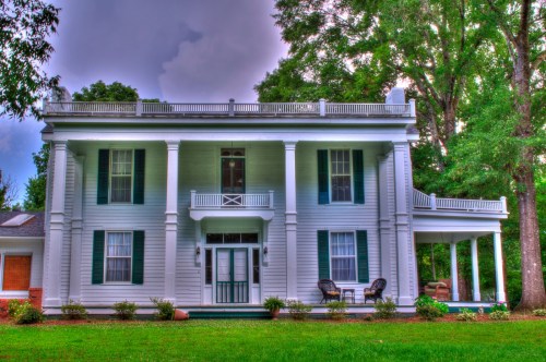 A large, elegant white house with green shutters, featuring a balcony and surrounded by lush trees and grass.