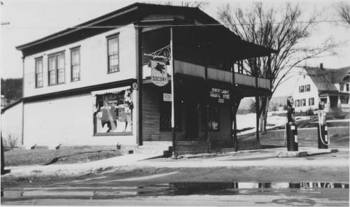 Historic black-and-white photo of a two-story building with a sign, gas pumps, and nearby trees.