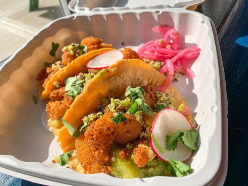 Three crispy shrimp tacos topped with cilantro, radish, and pickled onions, served in a takeout container.