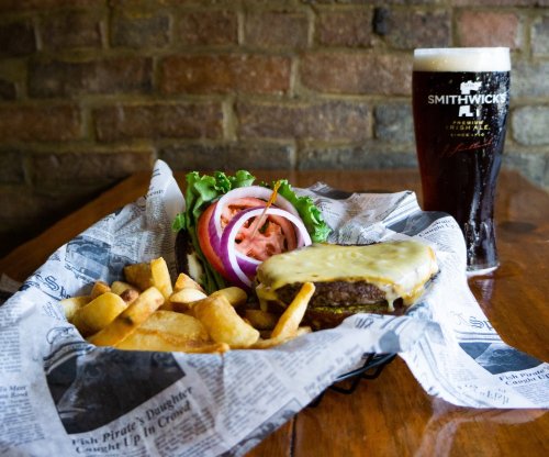 A cheeseburger with melted cheese, lettuce, tomato, and onion, served with fries and a pint of dark beer on a wooden table.