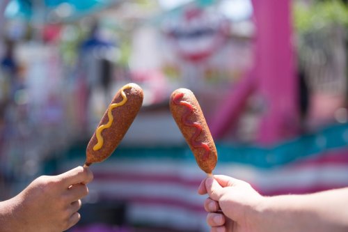 Two hands hold corn dogs topped with mustard and ketchup against a colorful fair background.