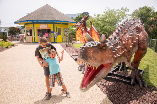 Two children pose playfully in front of a large dinosaur statue at a colorful attraction.