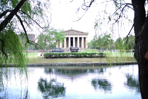 A classical building with columns reflected in a calm pond, surrounded by trees and greenery.