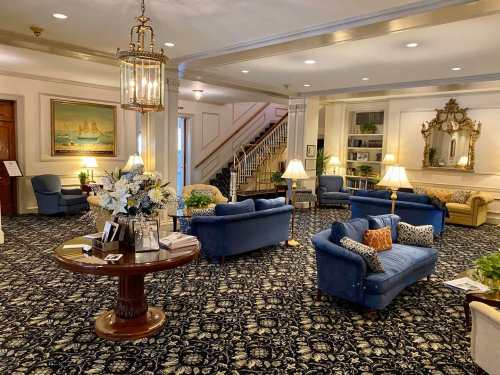 A cozy lobby with blue sofas, a decorative table, and elegant lighting, featuring a staircase and floral arrangements.