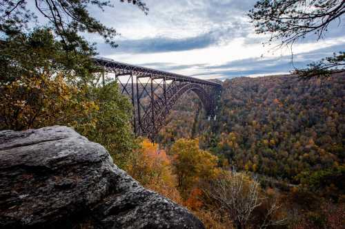 A towering bridge spans a colorful autumn valley, surrounded by trees and rocky terrain under a cloudy sky.