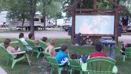 A group of children and adults sitting on lawn chairs watching a movie on an outdoor screen at a campsite.