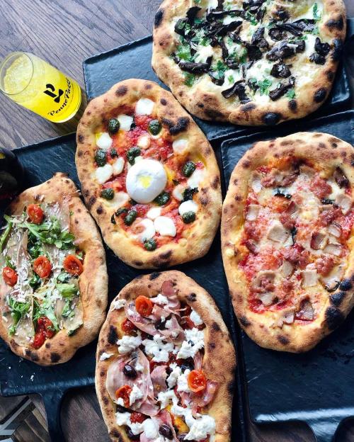 A variety of pizzas with different toppings on black plates, alongside a drink, arranged on a wooden table.