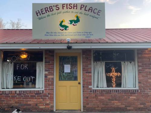 A brick building with a sign reading "Herb's Fish Place" and a window display saying "Open for Takeout."