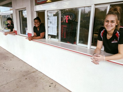 Three smiling employees lean out of a service window, welcoming customers at a food establishment.