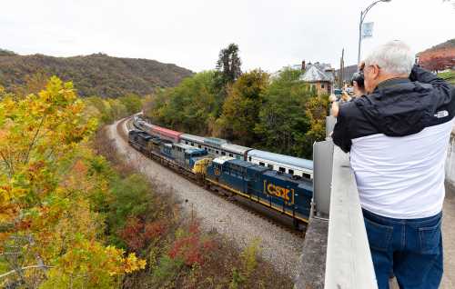 A person photographs a train passing through a scenic landscape with autumn foliage and hills in the background.