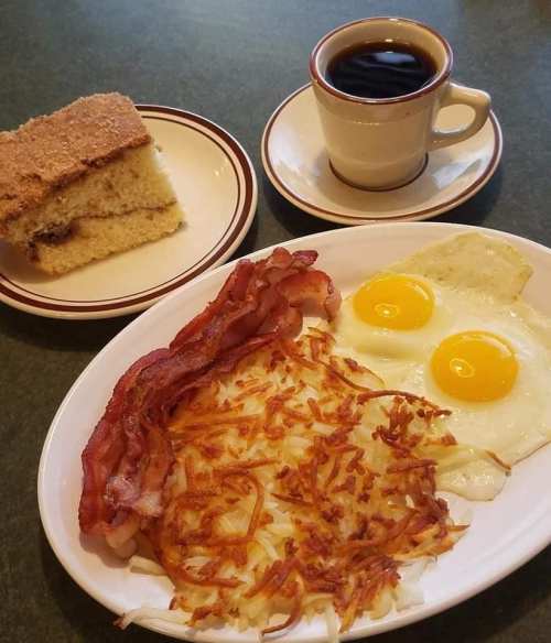 A plate with two fried eggs, crispy bacon, and hash browns, alongside a slice of cake and a cup of coffee.
