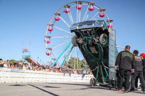 A truck performing a wheelie in front of a carnival with a ferris wheel and a crowd watching.