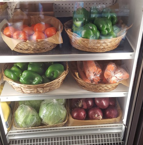 Baskets of fresh vegetables including tomatoes, bell peppers, carrots, lettuce, and onions in a refrigerated display.